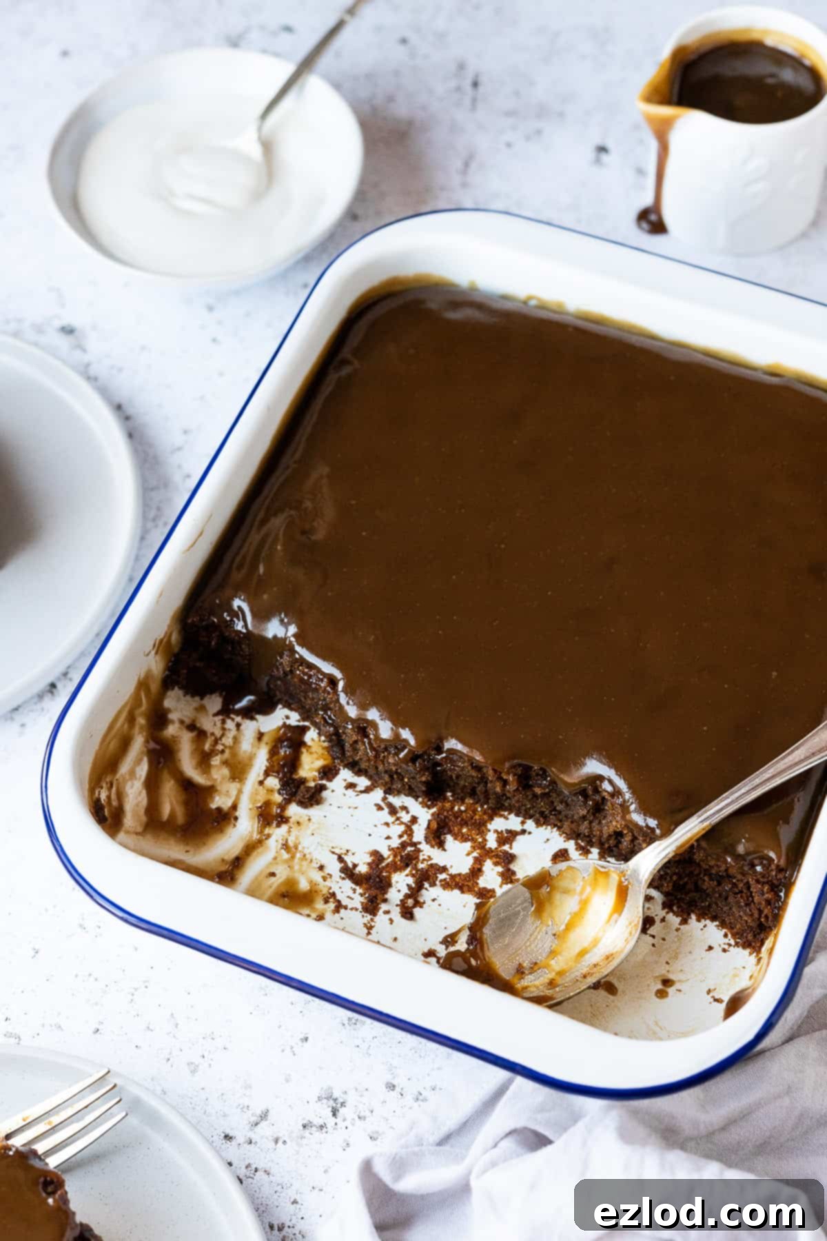 Sticky toffee pudding in a white enamel dish on a white background with a jug of sauce and a bowl of cream.