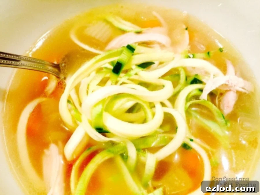 A steaming bowl of 21 Day Fix Chicken Zoodle Soup with shredded chicken, carrots, and celery, garnished with fresh herbs.