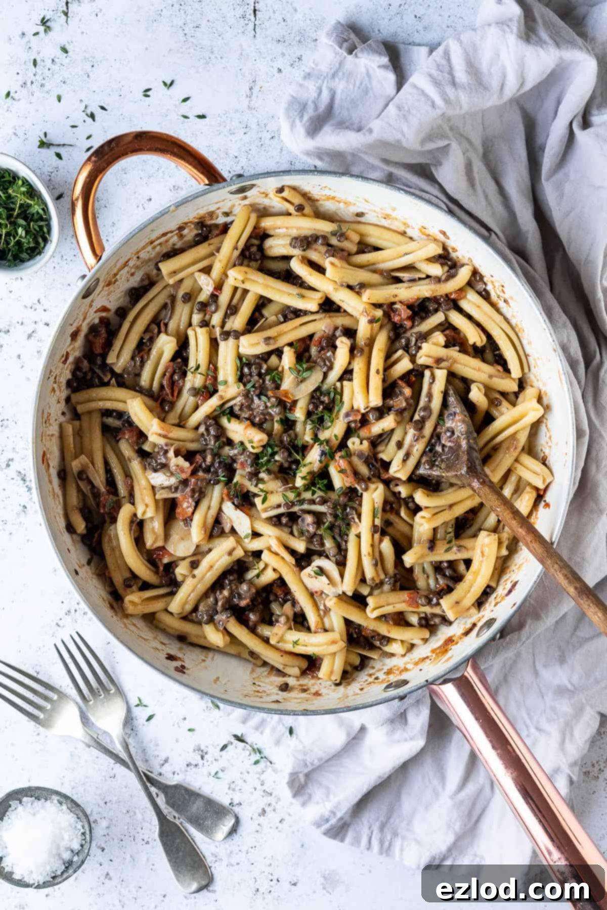 A white pan brimming with sun-dried tomato puy lentil pasta, resting on a rustic white surface. A grey cloth, two forks, a small bowl of salt, and a sprig of fresh thyme leaves complete the inviting scene, highlighting the dish's vibrant appeal.