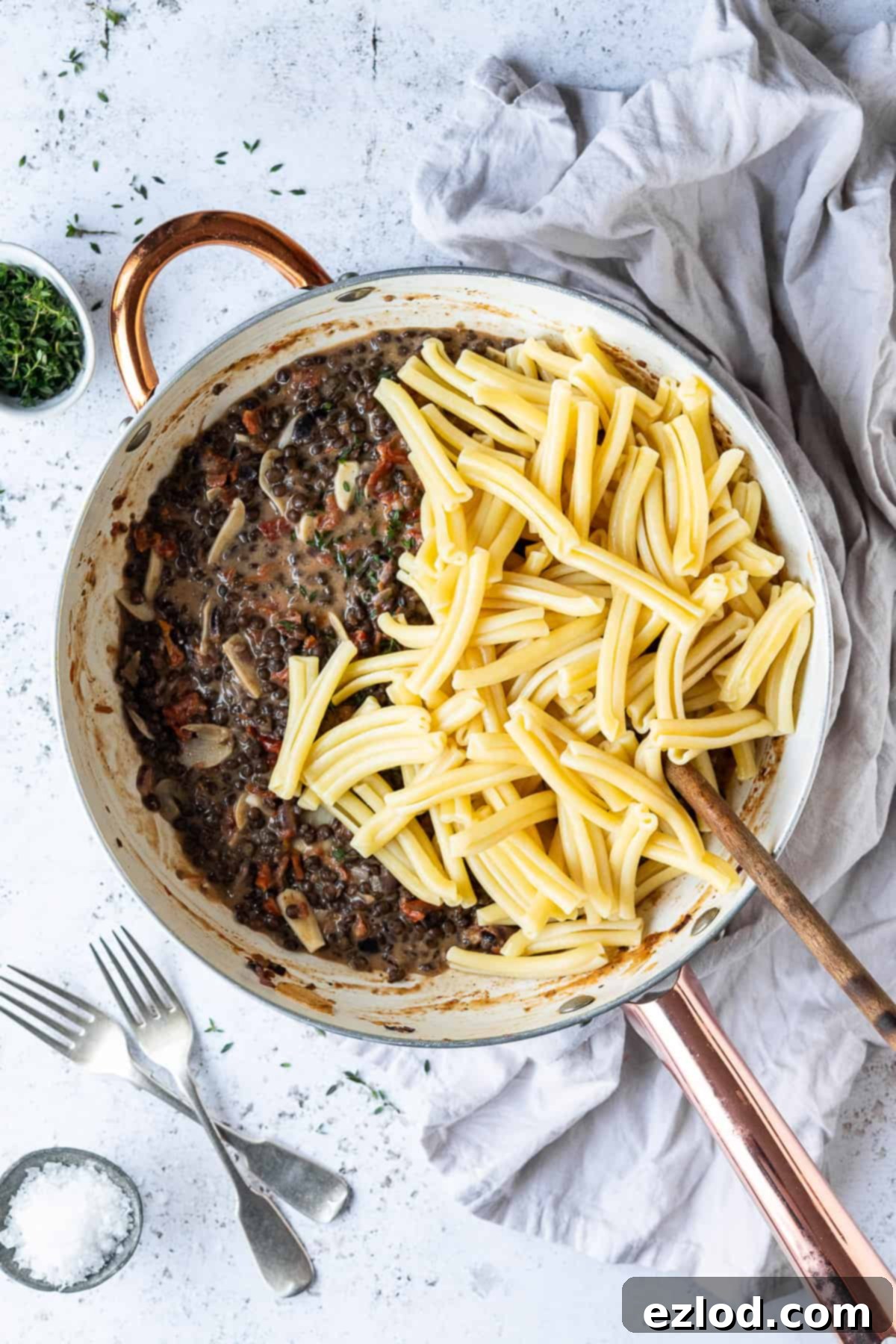 A large pan showcasing a rich sun-dried tomato puy lentil sauce, with cooked pasta shapes generously piled on top, ready to be mixed and served.