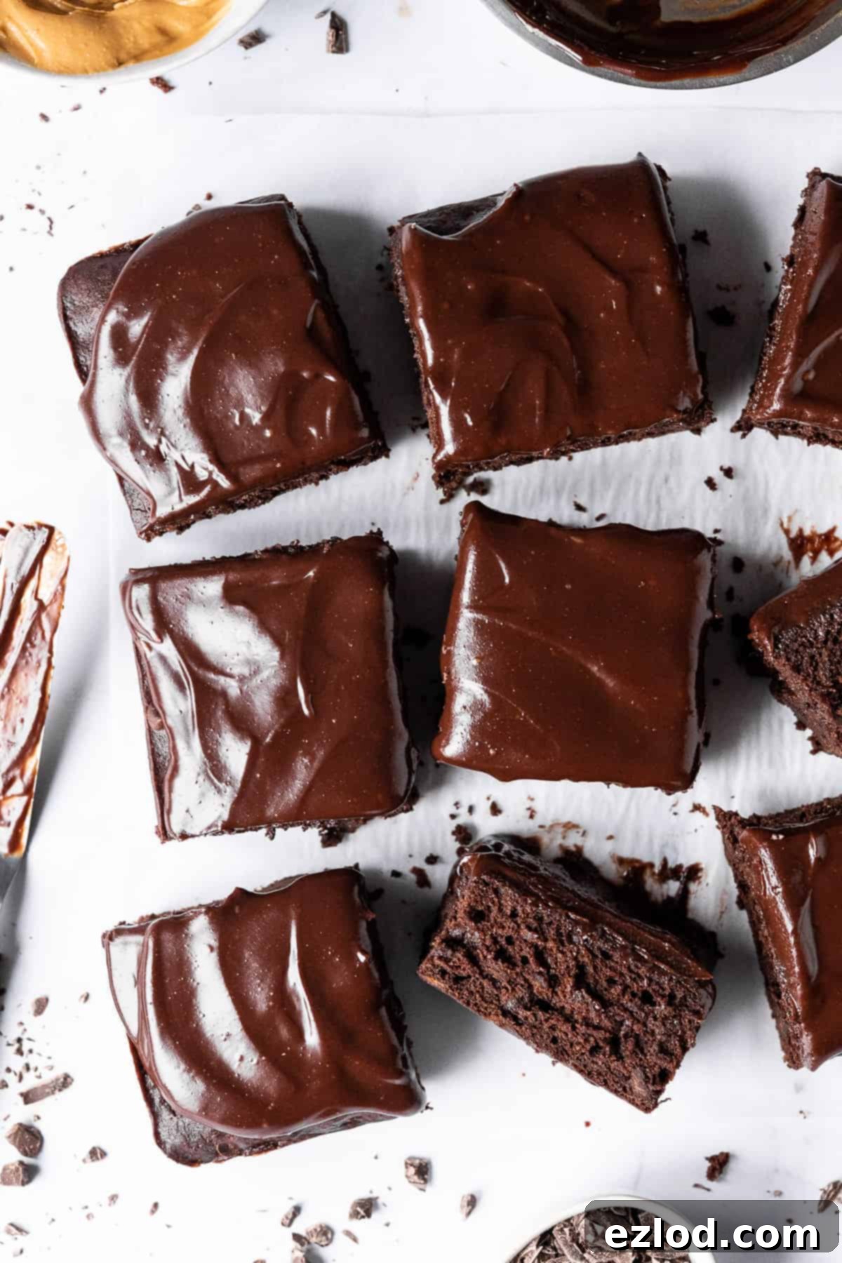 Squares of vegan chocolate banana cake on white baking parchment with a bowl of peanut butter, bowl of ganache, bowl of chopped chocolate and a ganache covered palette knife.