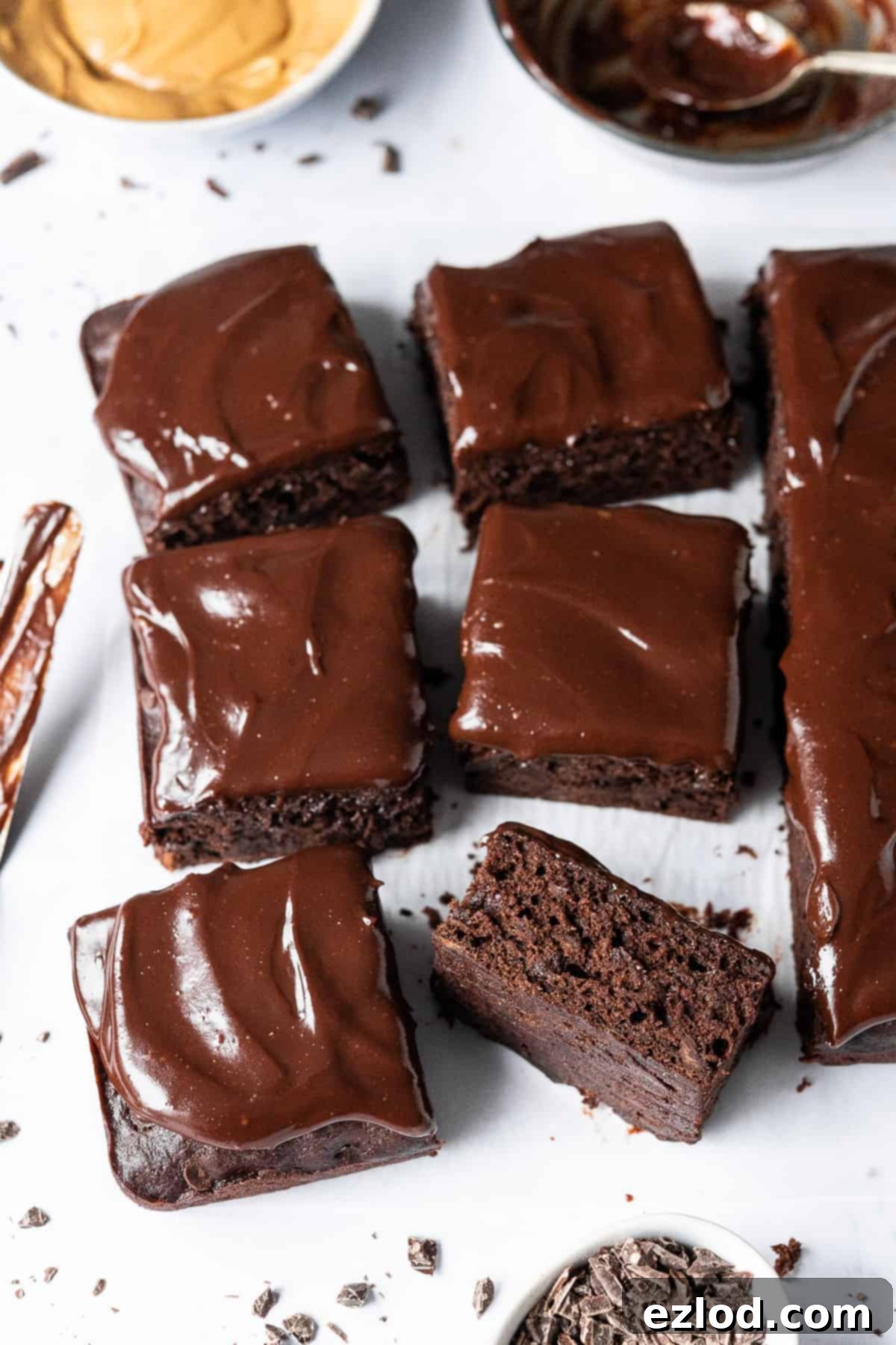 Squares of vegan chocolate banana cake on white baking parchment with a bowl of peanut butter and bowl of ganache in the background.