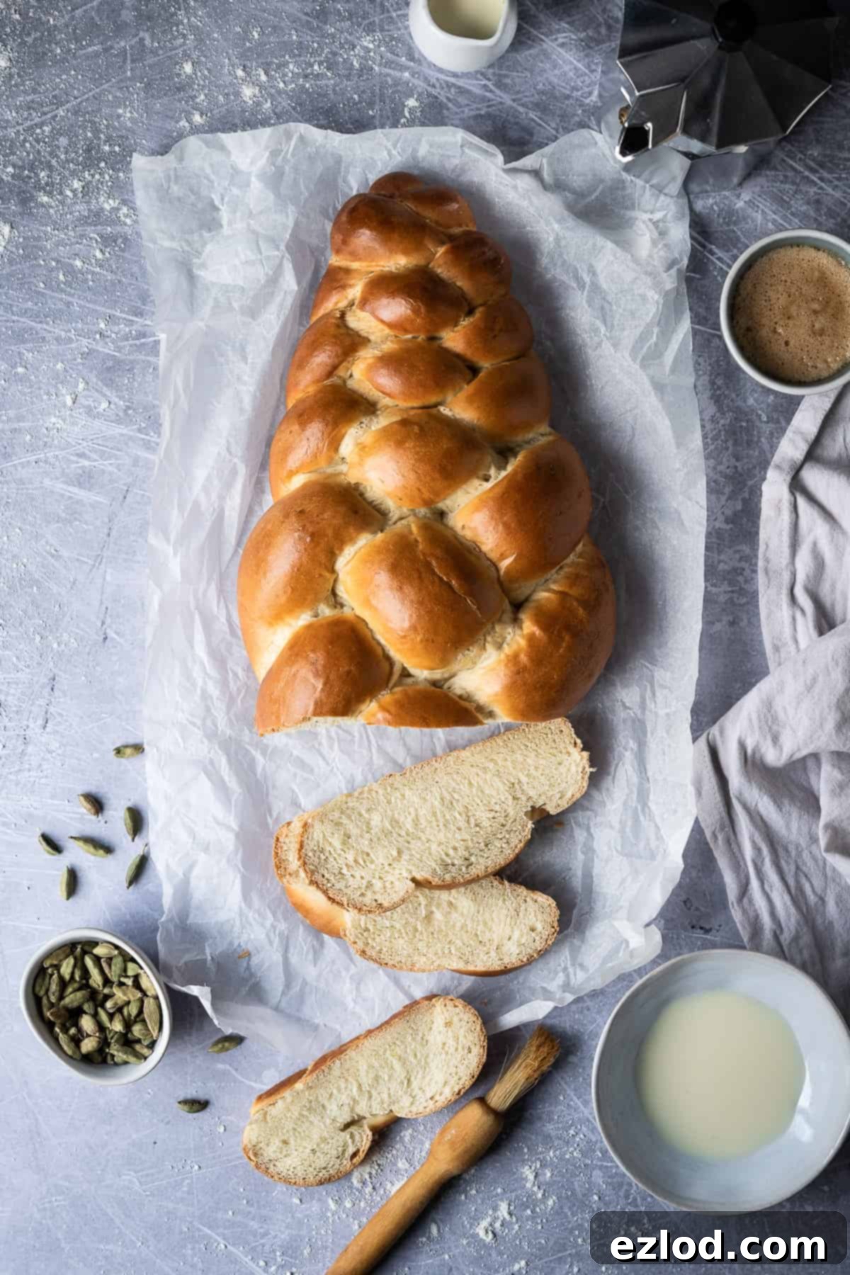 A beautifully sliced vegan pulla bread on parchment paper, showcasing its soft interior, surrounded by cardamom pods, a bowl of plant-based milk, a pastry brush, coffee accessories, and a cup of coffee. Perfect for a cozy breakfast or snack.