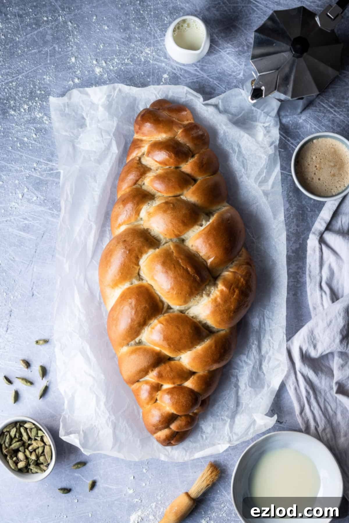 A golden-brown vegan pulla loaf, freshly baked and glistening from a milk wash, rests on parchment paper. Surrounded by cardamom pods, milk, coffee essentials, and a pastry brush, it exudes warmth and inviting aroma, ready to be enjoyed.