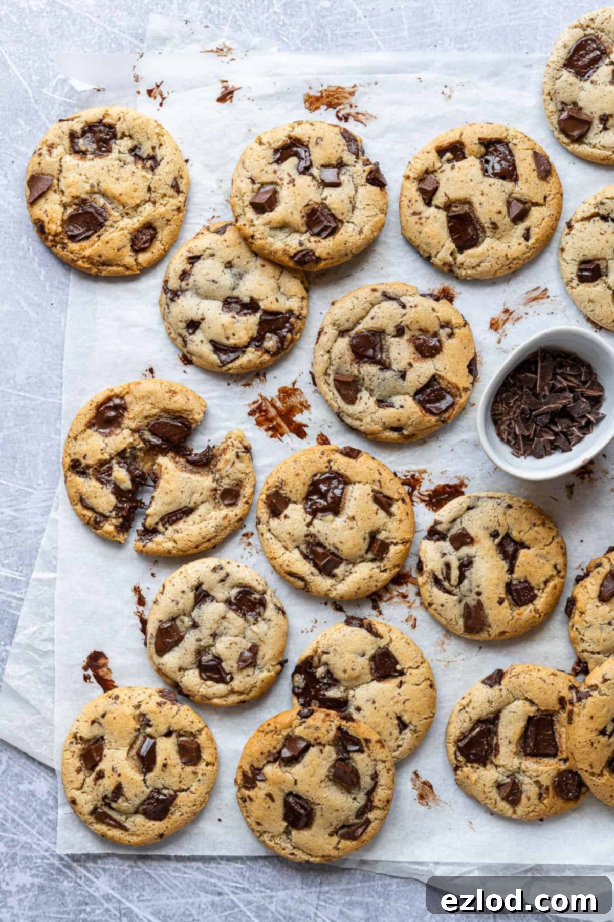 Vegan olive oil chocolate chip cookies and a bowl of chopped chocolate on a sheet of white baking parchment on a metal surface.