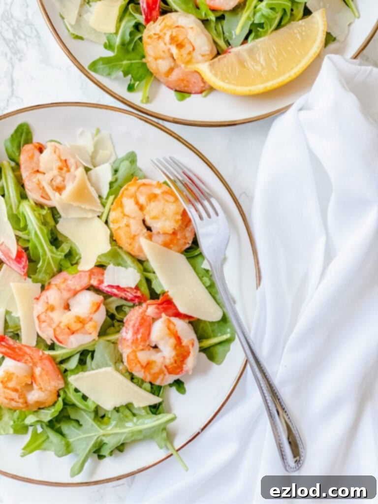 Two plates of arugula salad with shrimp and Parmesean on a white background with a fork and white napkin on the side.