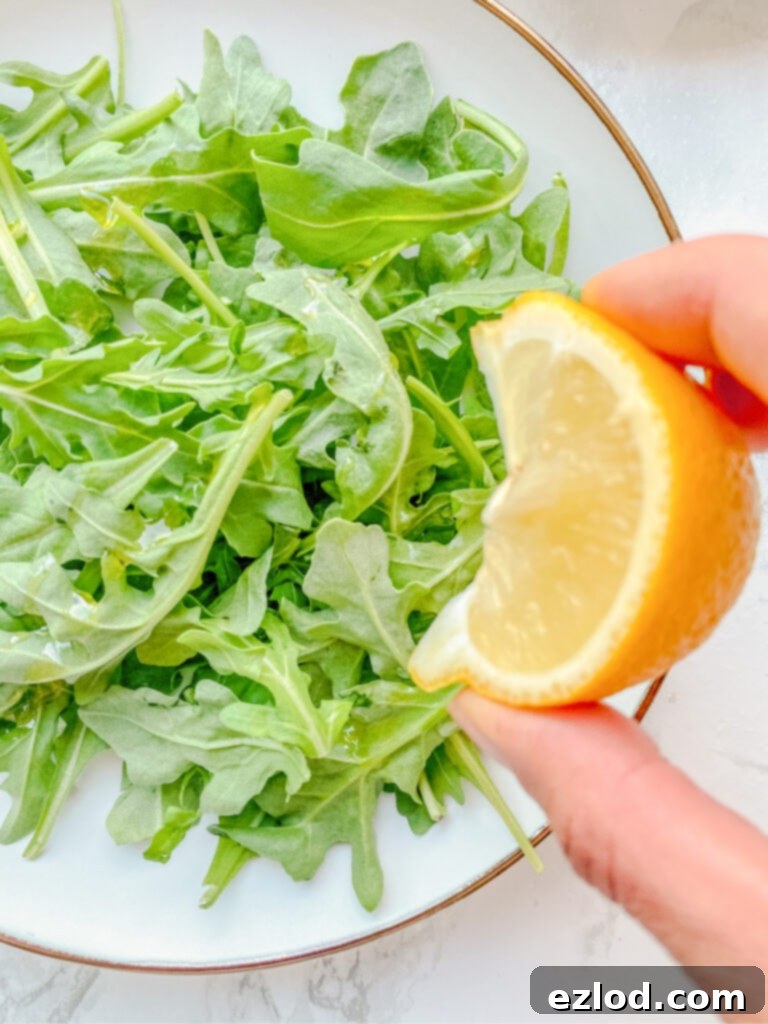 Close up photo of a hand squeezing a slice of lemon onto a bed of arugula on a white plate.