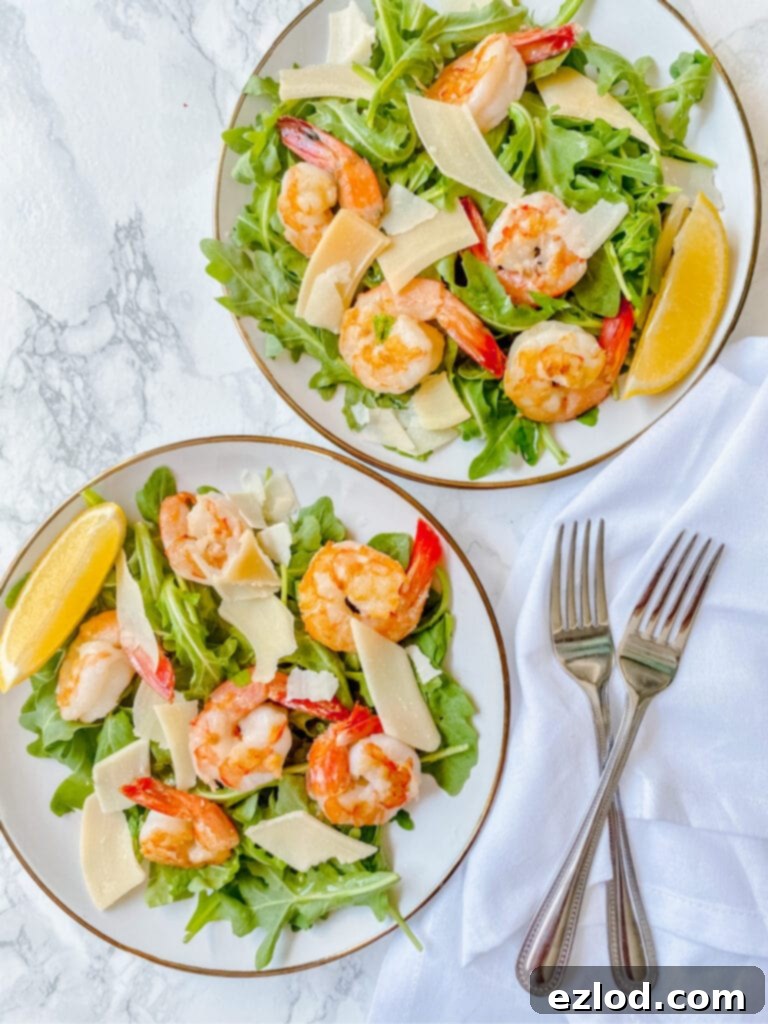 Overhead photo of two arugula salads topped with shrimp, shaved parmesan cheese, and lemon slices. There are two forks forming an X on a white napkin off to the side.