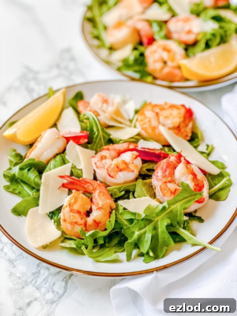 High angle photo of arugula salad with shrimp on a white plate. There is a second arugula salad recipe in the background, out of focus.