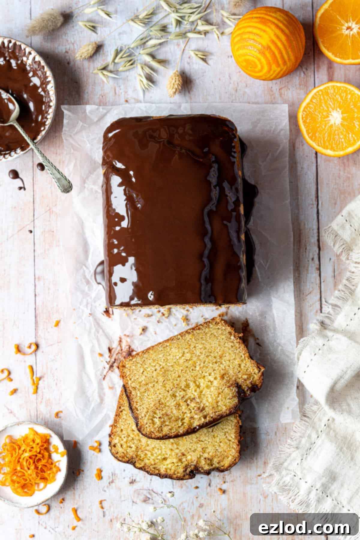 A beautifully presented vegan orange loaf cake on a rustic wooden table, surrounded by fresh oranges, a bowl of rich chocolate glaze, fragrant orange zest, and a soft linen cloth.