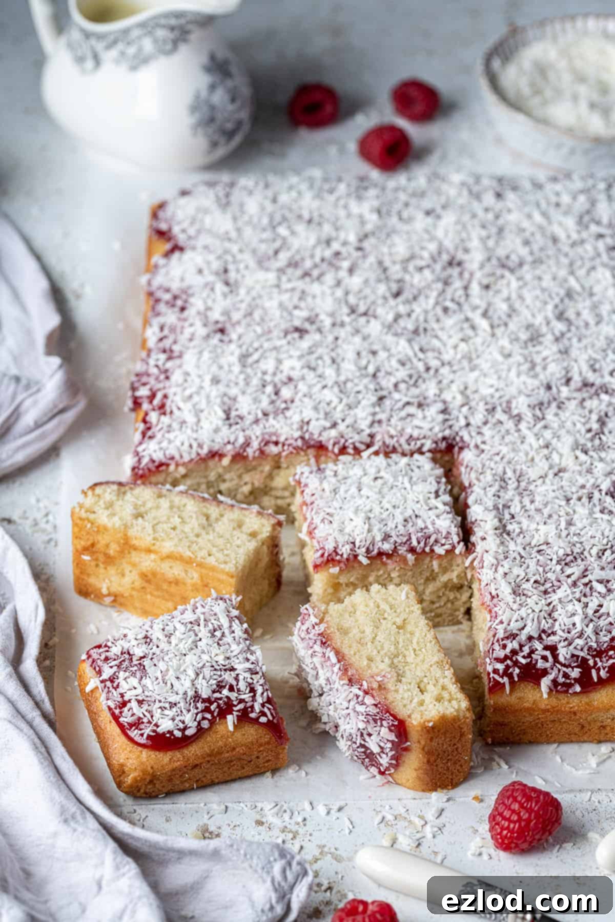 Vegan jam and coconut sponge cake on a white table with raspberries, a grey cloth, jug of custard and bowl of coconut.