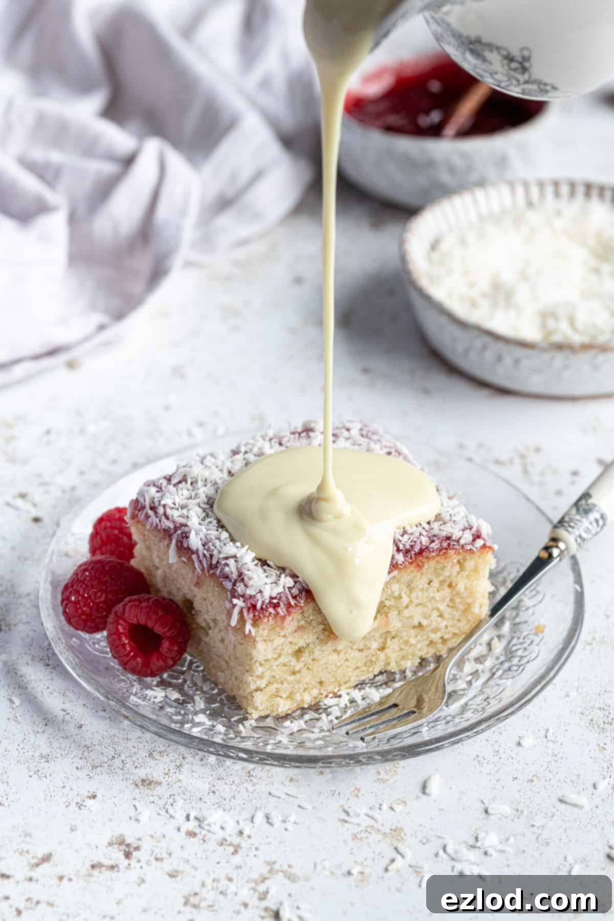 Rich vegan vanilla custard being generously poured from a white jug over a slice of cake on a plate.