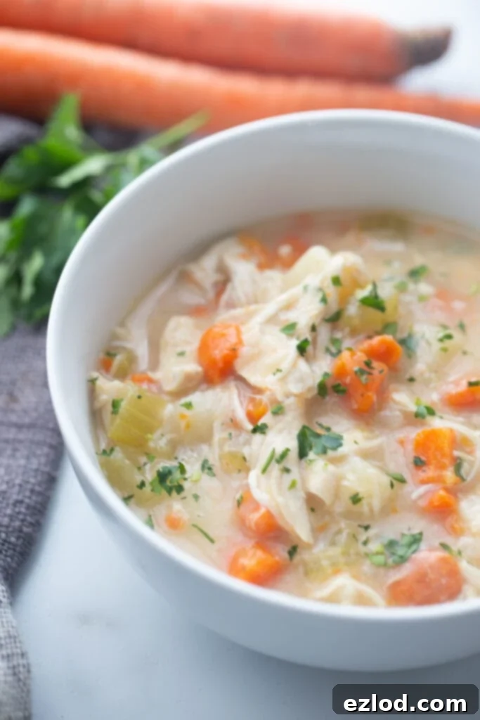 A steaming white bowl of healthy Instant Pot chicken pot pie soup, garnished with fresh parsley, sits on a white surface with blurred carrots in the background.