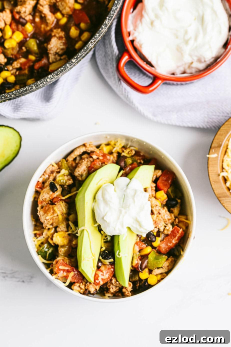 A white marble background displaying a small bowl of taco skillet topped with avocado and Greek yogurt, with the remaining skillet and a bowl of plain Greek yogurt nearby for serving.