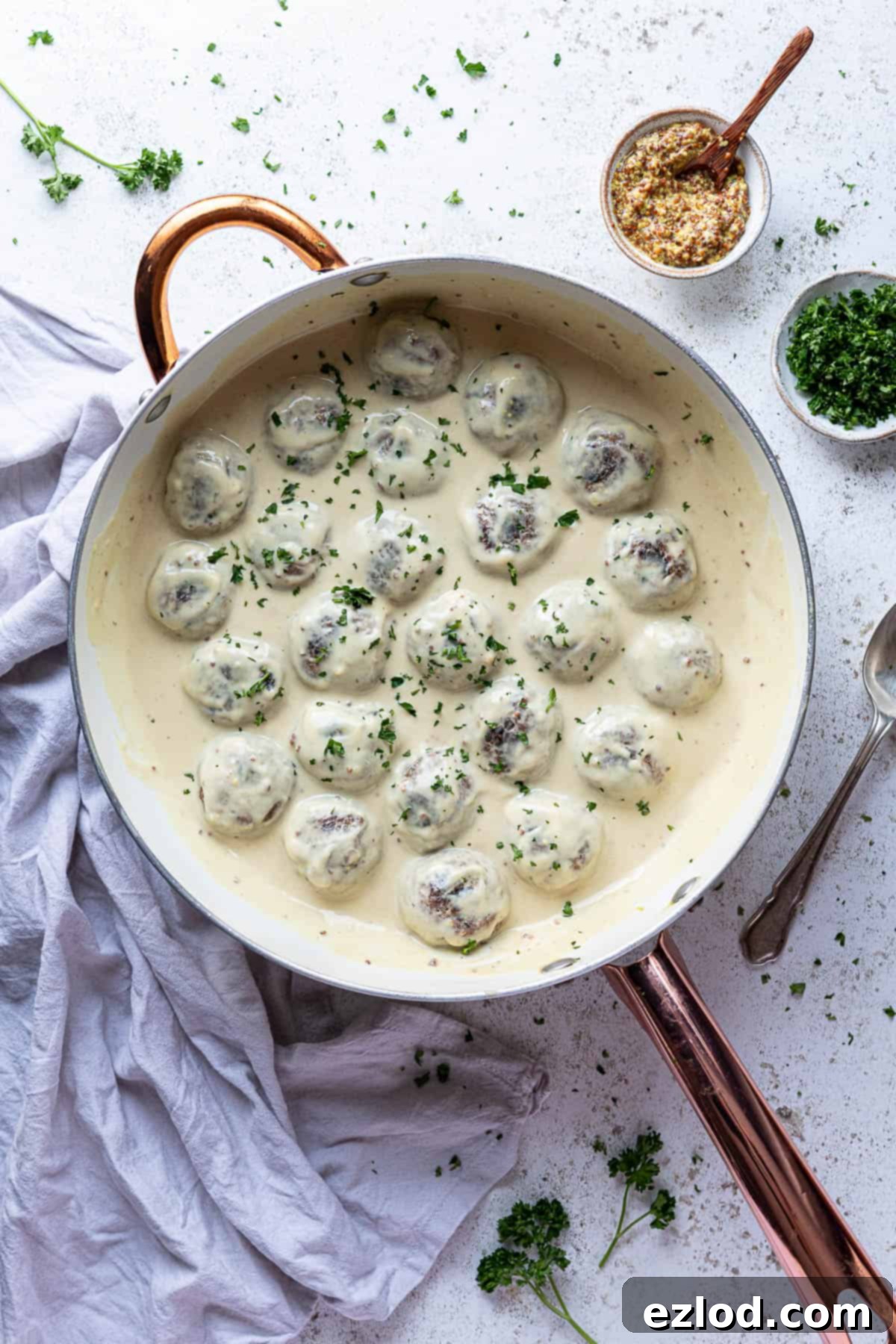 Vegan Swedish meatballs in a creamy mustard sauce in a white saucepan with a bowl of mustard and a bowl of chopped parsley.