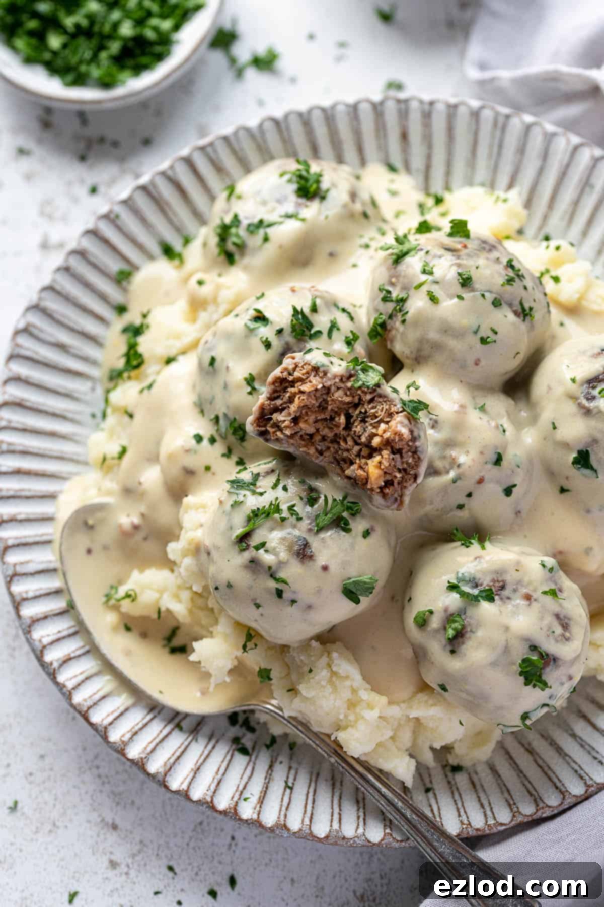 Vegan Swedish meatballs on top of mashed potato on a white plate, one with a bite taken out of it.