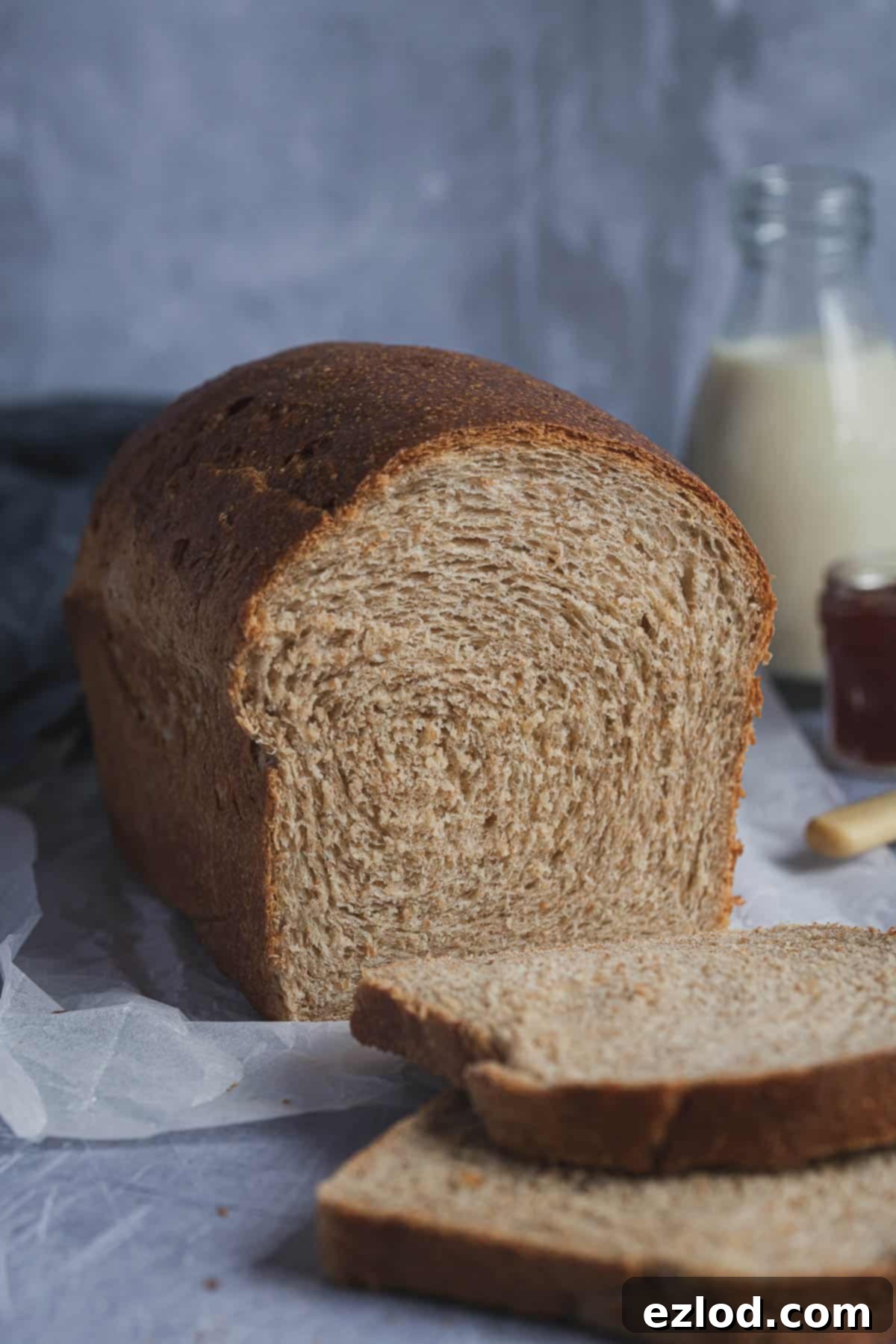 A beautifully baked loaf of vegan wholemeal sandwich bread, with two slices cut, revealing its soft texture. In the background, a bottle of plant-based milk and a jar of homemade jam add to the cozy scene.