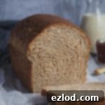 A loaf of vegan wholemeal sandwich bread with two slices cut off and a bottle of milk and jar of jam in the background.