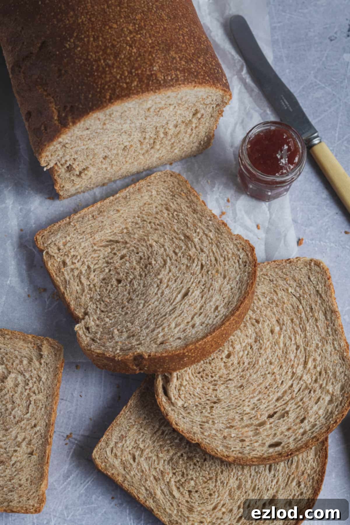 Slices of freshly baked wholemeal sandwich bread neatly arranged on white baking parchment, with a rustic jar of jam and a bread knife nearby, inviting a delicious breakfast.