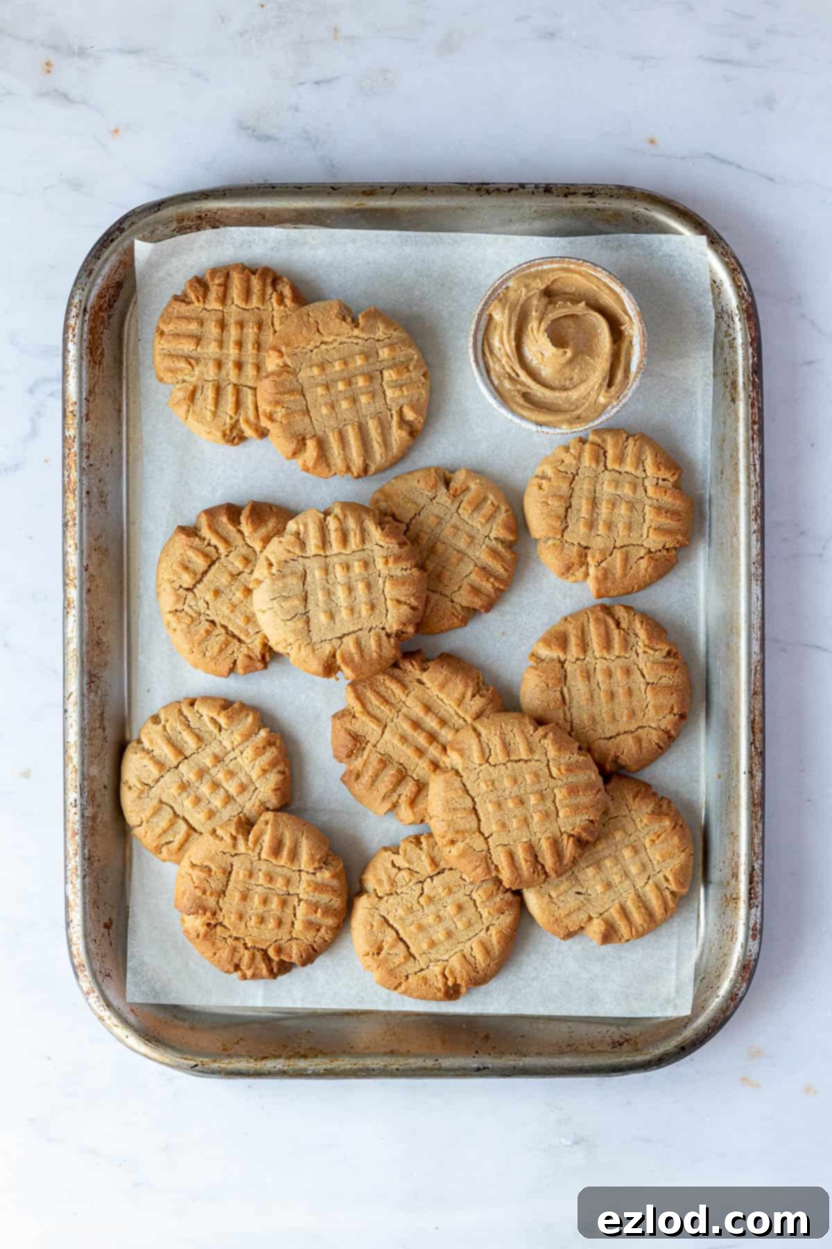 A batch of unbaked vegan peanut butter cookie dough balls on a metal baking sheet, next to a bowl of creamy peanut butter.