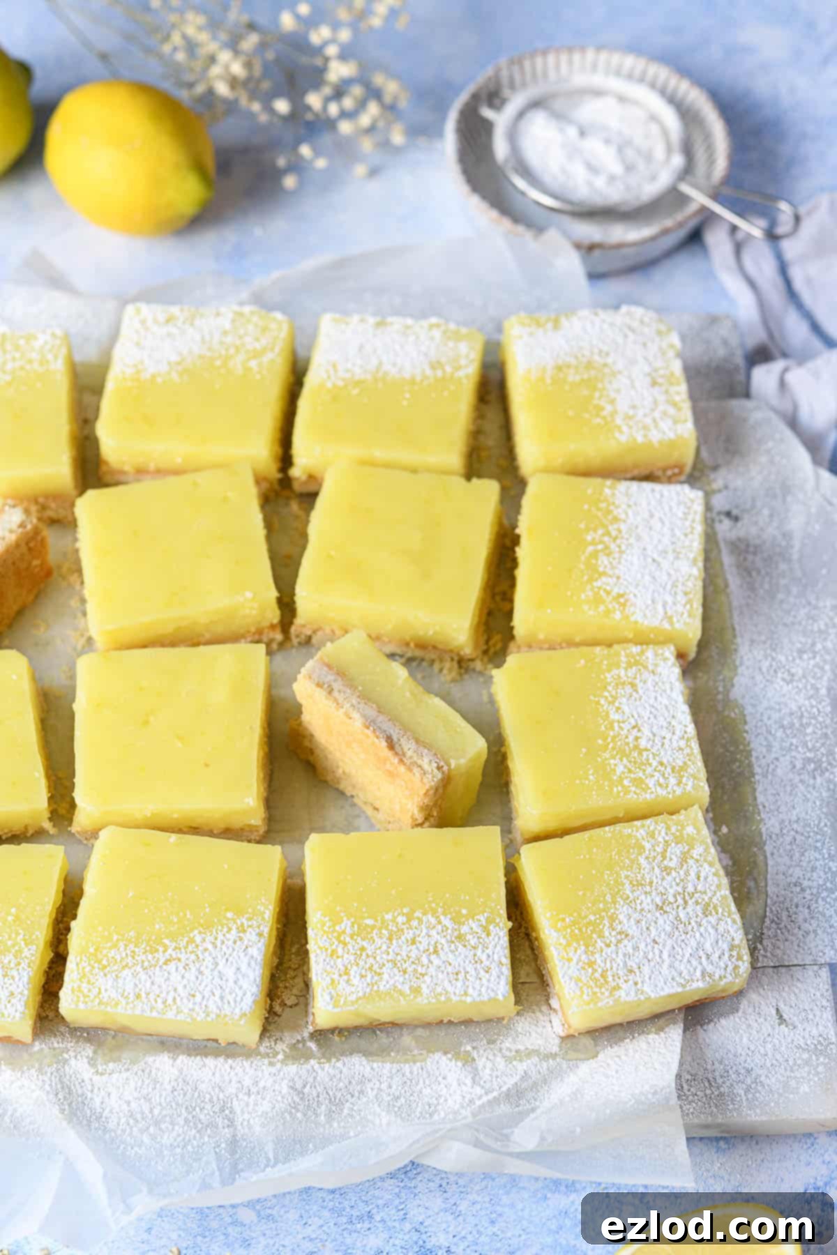 Freshly baked lemon bars resting on white baking parchment atop a vibrant blue surface, awaiting slicing.