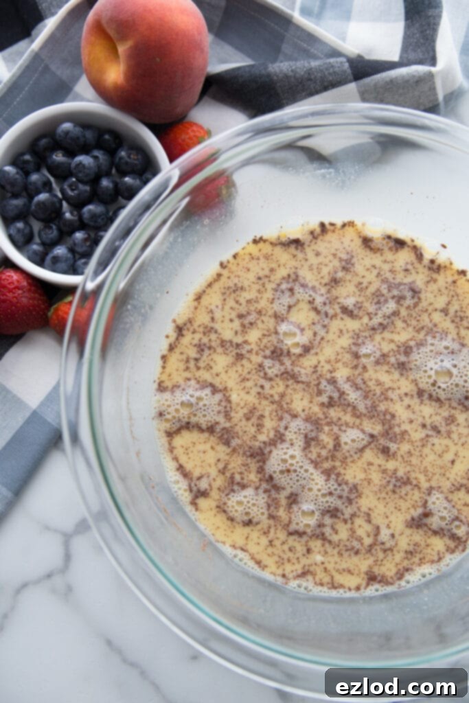A glass bowl of french toast custard with cinnamon sitting next to a bowl of blueberries and a peach