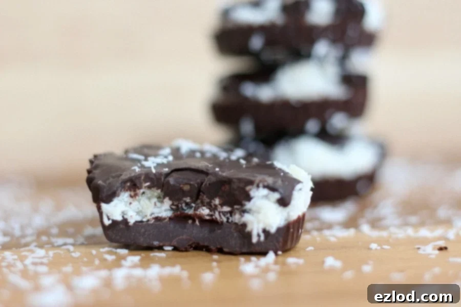 Close up side view of a homemade chocolate coconut candy cup with a bite taken out. Out of focus in the background is a stack of 3 coconut cups. There is coconut sprinkled on the wooden tabletop.