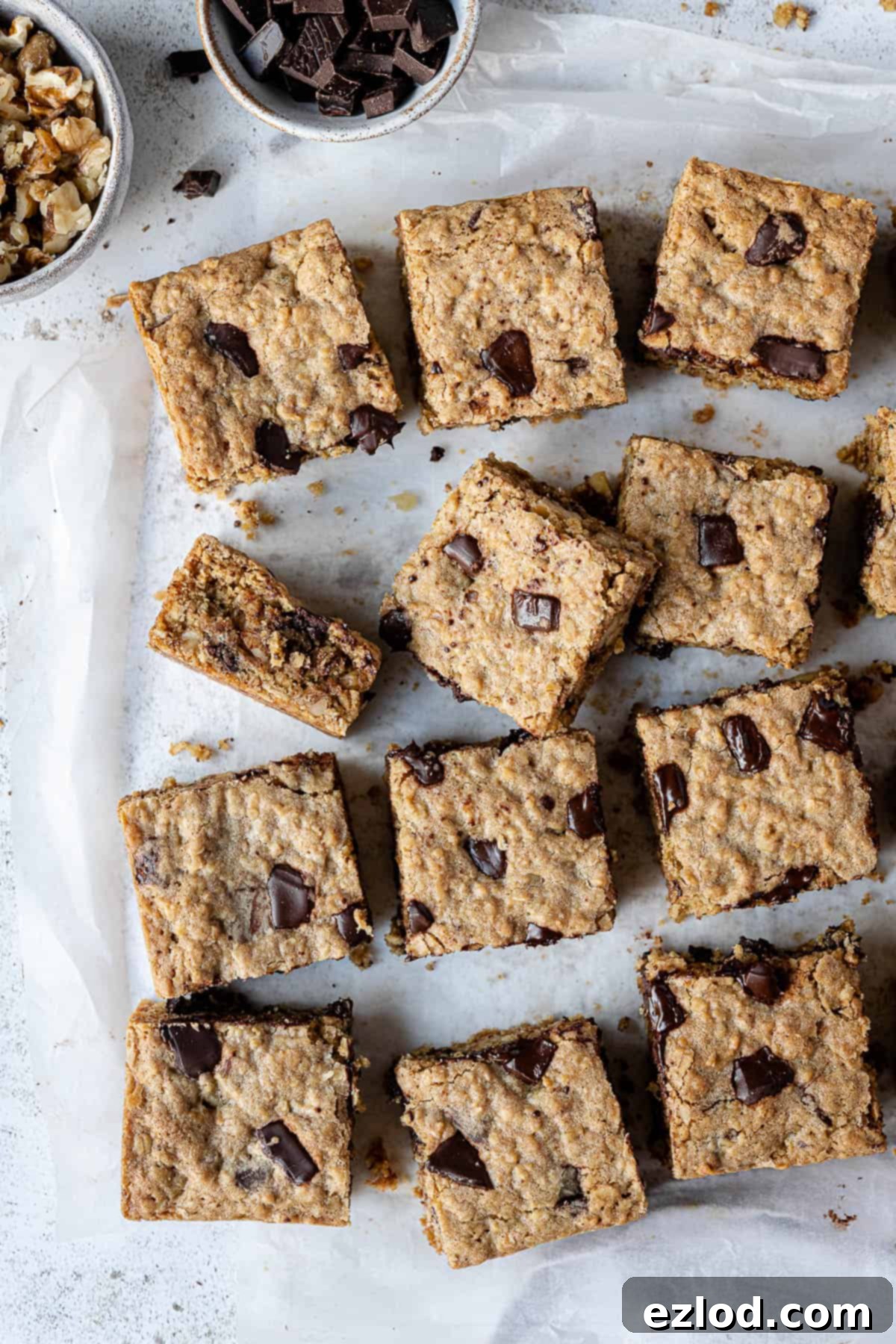 Close-up of perfectly baked vegan oatmeal cookie bars, studded with chocolate and walnuts, resting on baking parchment next to bowls of their key ingredients.