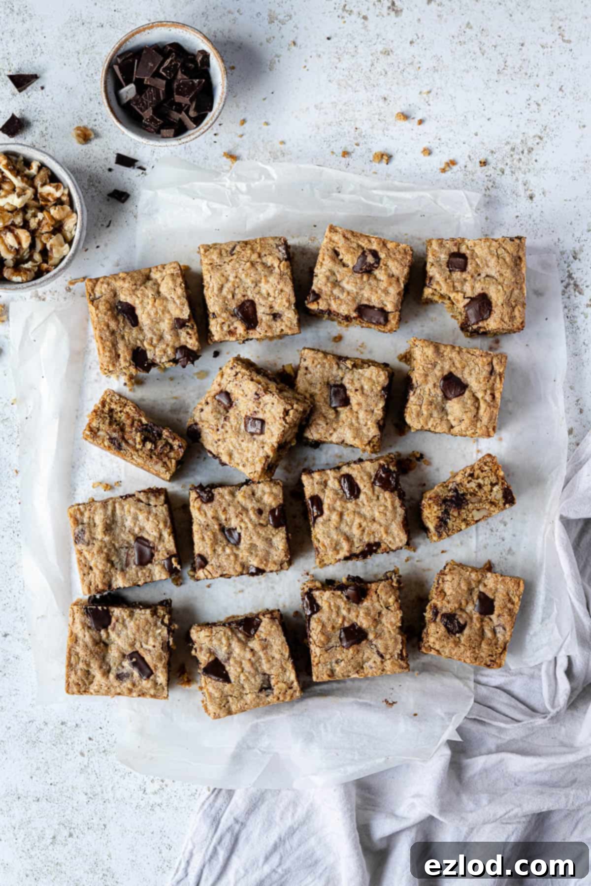 A beautifully arranged image featuring vegan oatmeal cookie bars on baking parchment, accompanied by bowls of chocolate and walnuts, and a stylish grey cloth – perfect for Pinterest.