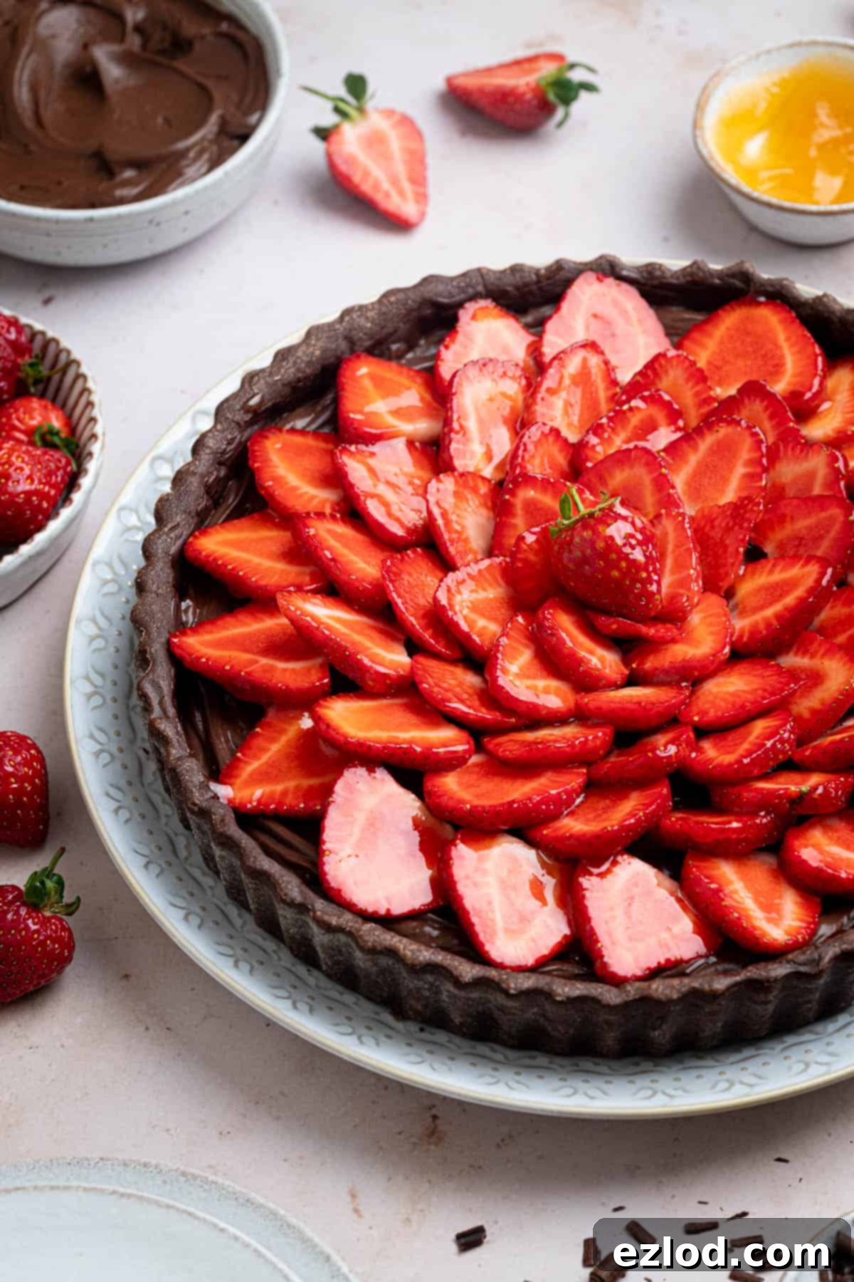 A beautifully presented slice of vegan chocolate strawberry tart on a white plate, accompanied by bowls of creamy chocolate pastry cream, fresh strawberries, and glistening apricot jam.