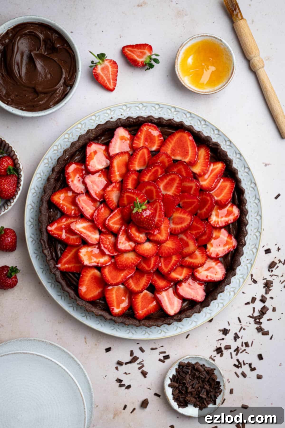 A beautifully composed flat lay of a full vegan chocolate strawberry tart on a white serving plate, surrounded by small bowls containing fresh strawberries, creamy chocolate pastry cream, delicate chocolate shavings, and shimmering apricot jam glaze.