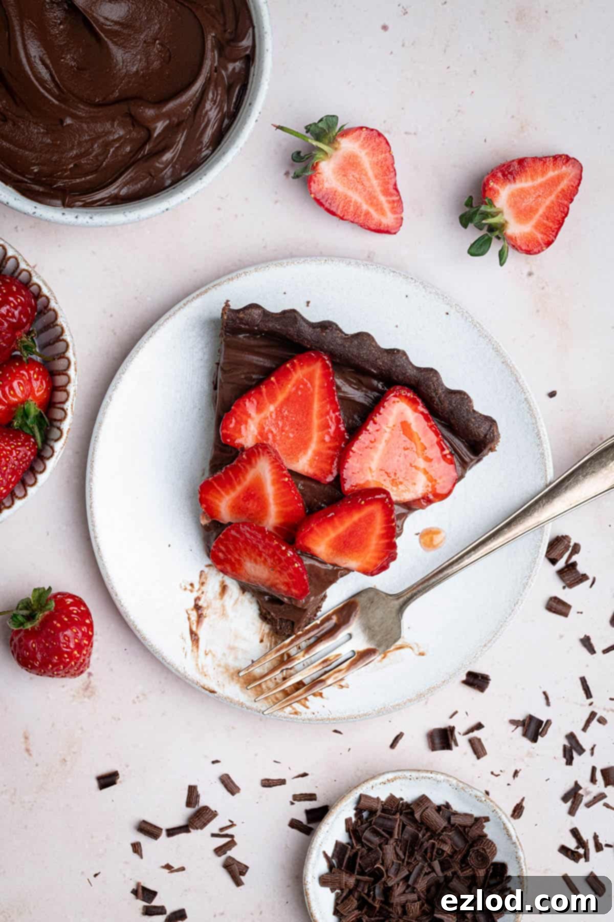 A close-up shot of a single slice of vegan chocolate strawberry tart on a white plate, with a fork having removed a small portion, highlighting the layers of crisp chocolate pastry, creamy chocolate filling, and fresh strawberries.