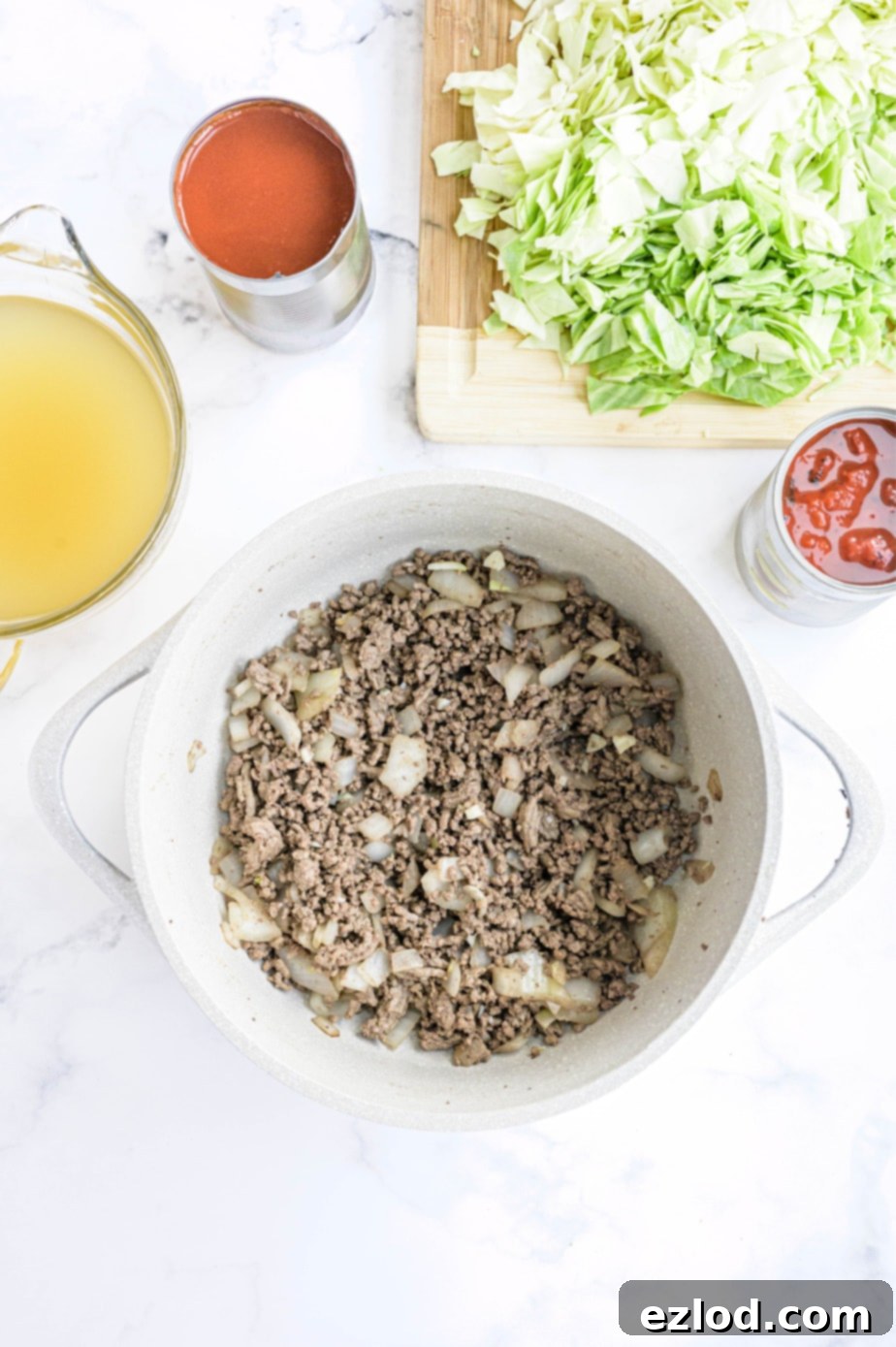 Fresh ingredients for unstuffed cabbage roll soup including ground meat, chopped cabbage, onions, and tomatoes are laid out on a white surface.