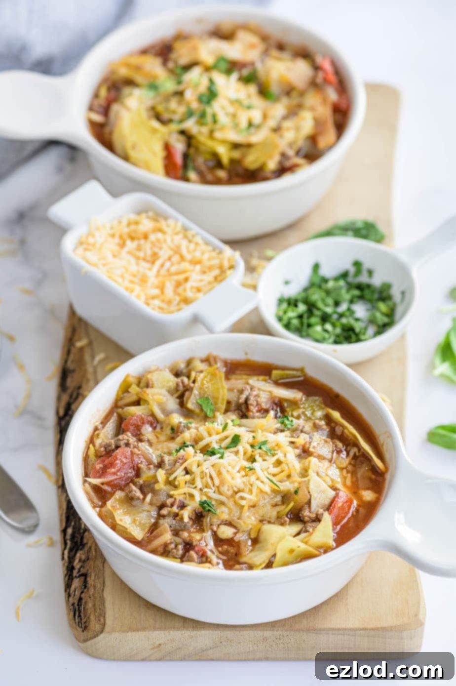 Two bowls of unstuffed cabbage roll soup are placed next to small bowls of cheese and parsley for garnish.