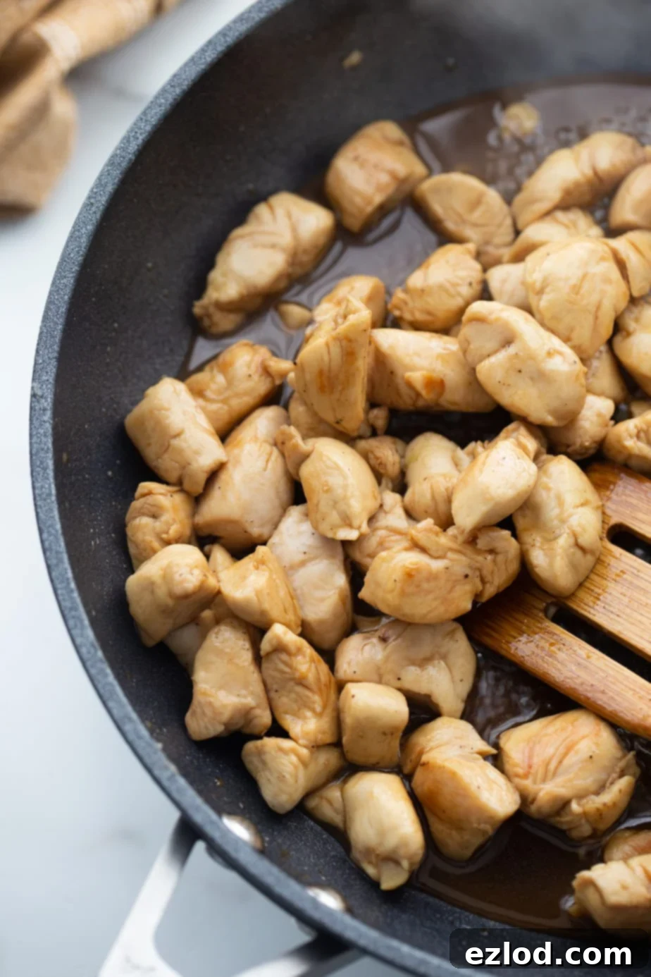 Browned chicken is being stirred by a wooden spoon in a black skillet, coated in a rich sauce.