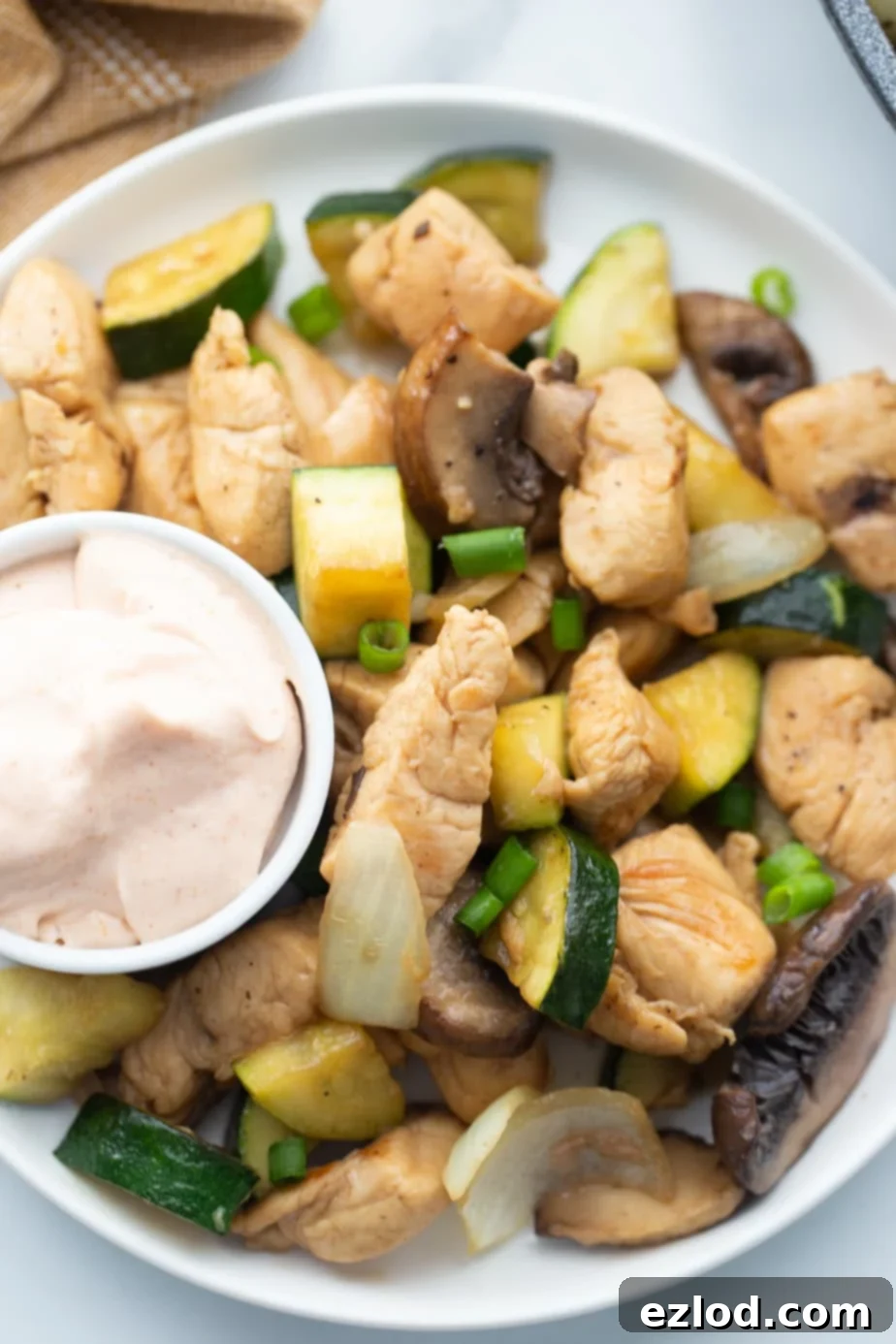 A small cup of creamy yum yum sauce sits on a white plate next to a generous serving of cooked hibachi chicken and stir-fried vegetables.