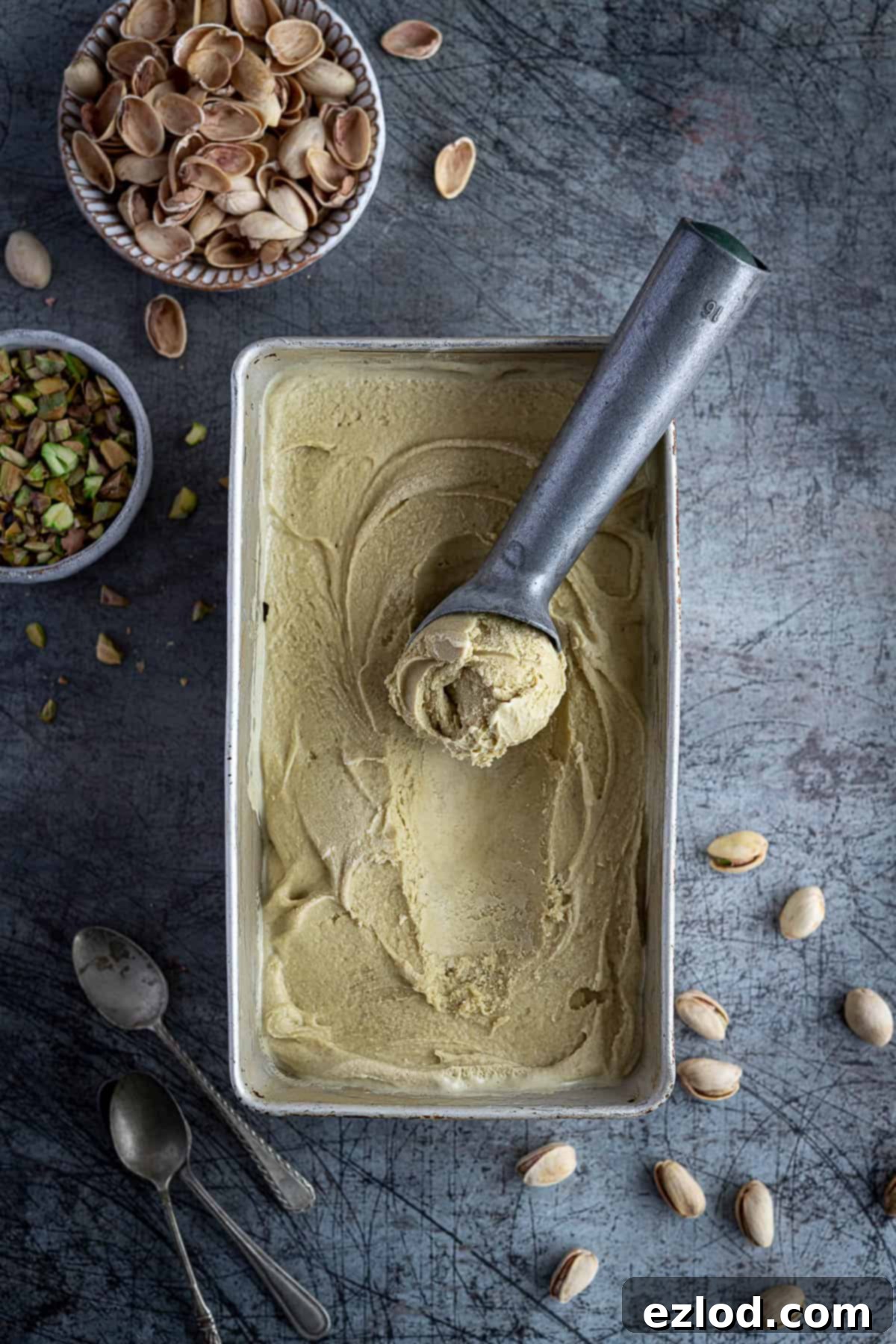 Vegan pistachio ice cream in a metal loaf tin with an ice cream scoop, bowl of chopped pistachios and a bowl of pistachio shells.