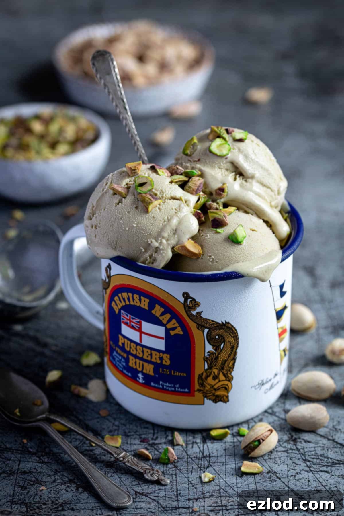 Scoops of vegan pistachio ice cream in an enamel mug with a bowl of chopped pistachios and a bowl of pistachio shells.