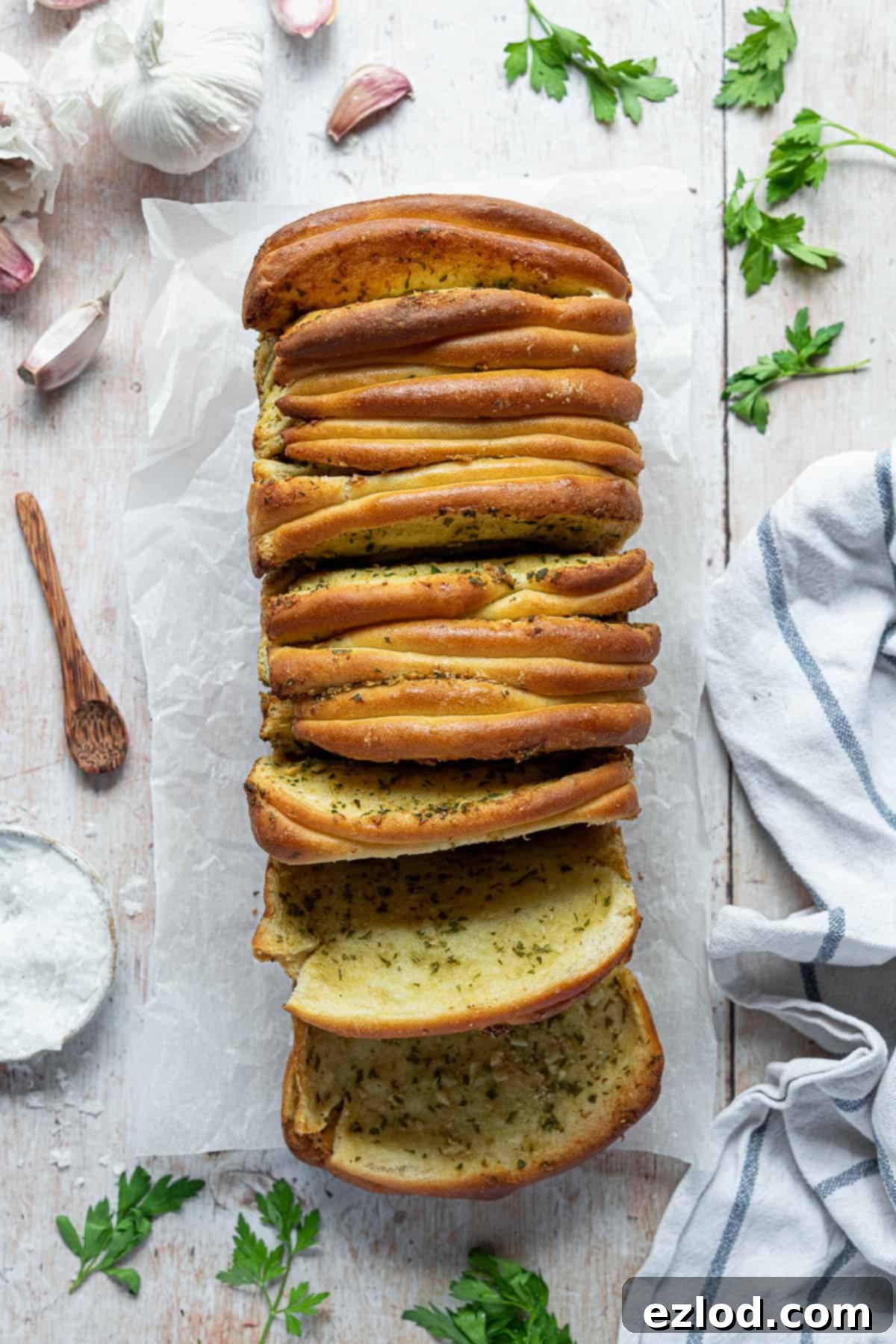 Freshly baked vegan garlic pull apart bread on a rustic wooden table, surrounded by whole garlic cloves, fresh parsley, a small bowl of sea salt, and a soft tea towel. The bread has a golden-brown crust and a visible soft texture.