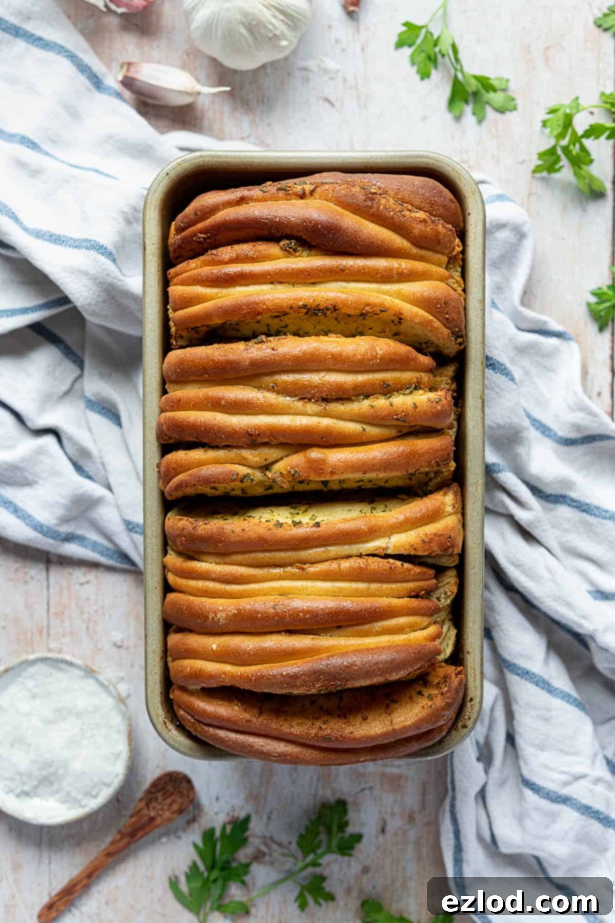 A beautifully arranged vegan garlic pull apart bread in a loaf tin, set on a wooden table. Accompanying items include a blue and white tea towel, fresh garlic cloves, vibrant parsley, and a small ceramic bowl of salt, creating an inviting rustic scene.