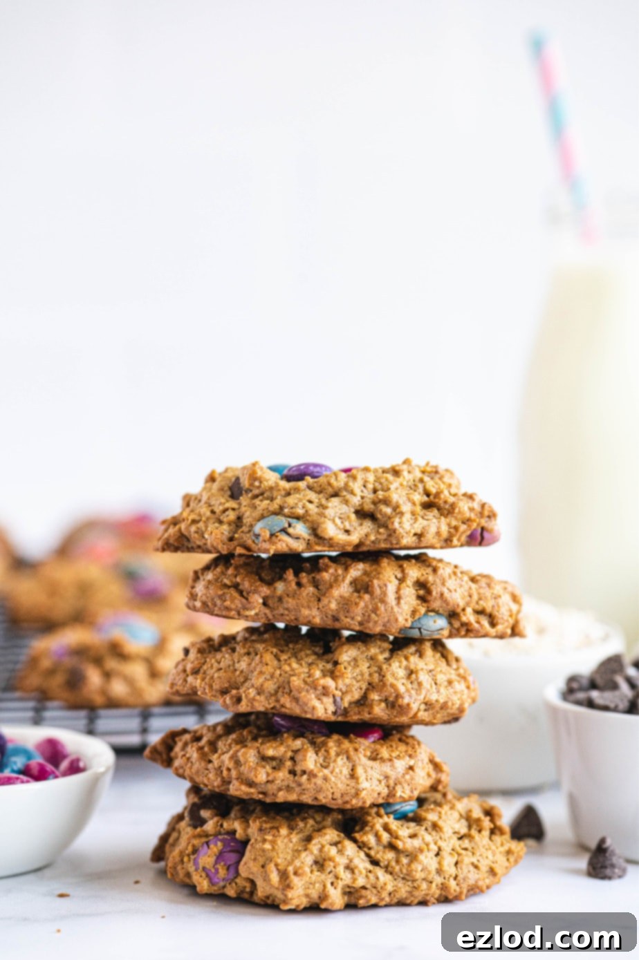 A stack of golden brown monster cookies, studded with colorful dairy-free chocolate candies, is presented on a clean white surface, invitingly ready to be enjoyed.