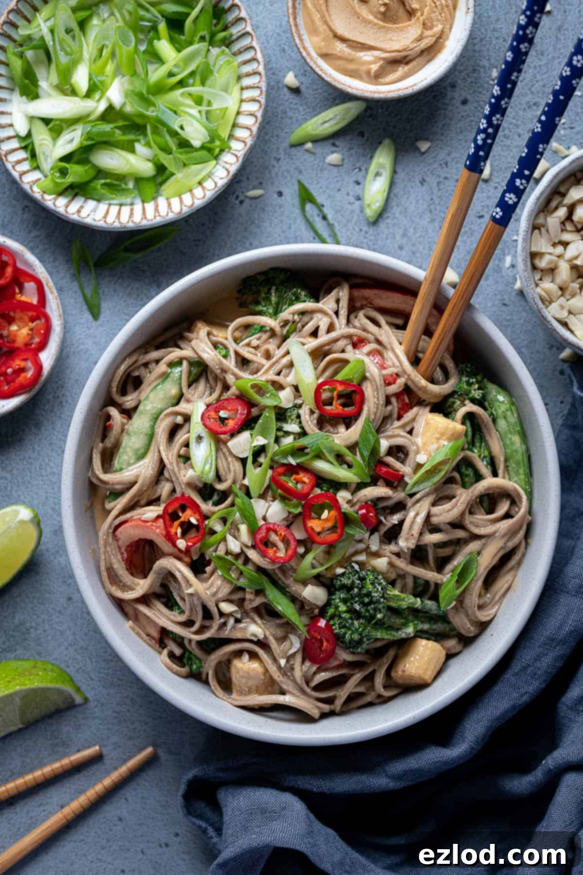 Creamy Peanut Noodles: A fast, flavourful, and satisfying vegan meal. A vibrant bowl of creamy peanut noodles topped with stir-fried vegetables, fresh spring onions, and a sprinkle of chopped peanuts. Chopsticks rest on the edge of the bowl, with smaller bowls of fresh chillies, lime wedges, and peanut butter in the background, highlighting the fresh ingredients.
