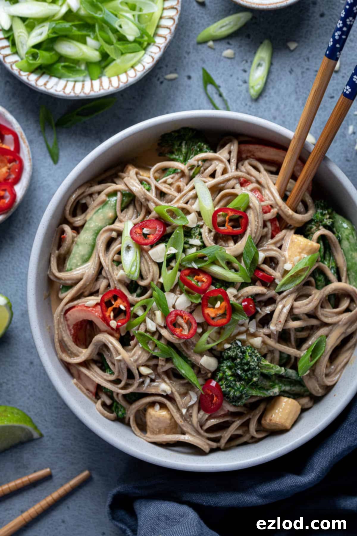 A delicious and colourful bowl of creamy peanut noodles with vegetables and garnishes. Close-up of a bowl filled with creamy peanut noodles, mixed with stir-fried green vegetables and topped with fresh sliced spring onions and vibrant red chillies, ready to be enjoyed with chopsticks.