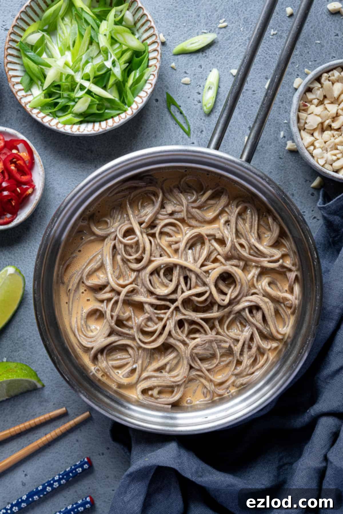 Creamy peanut noodles in a pan with garnishes. A large pan filled with creamy peanut noodles, beautifully coated in sauce. Around the pan are small bowls containing garnishes: chopped peanuts, sliced spring onions, fresh red chillies, and vibrant lime wedges, ready for customization.