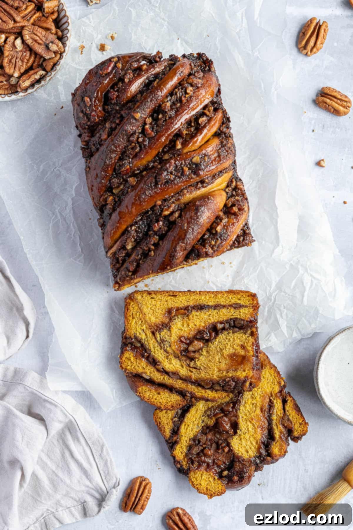 A loaf of pumpkin pecan babka with two slices cut off, with a bowl of pecans, a bowl of sugar syrup and a pastry brush.