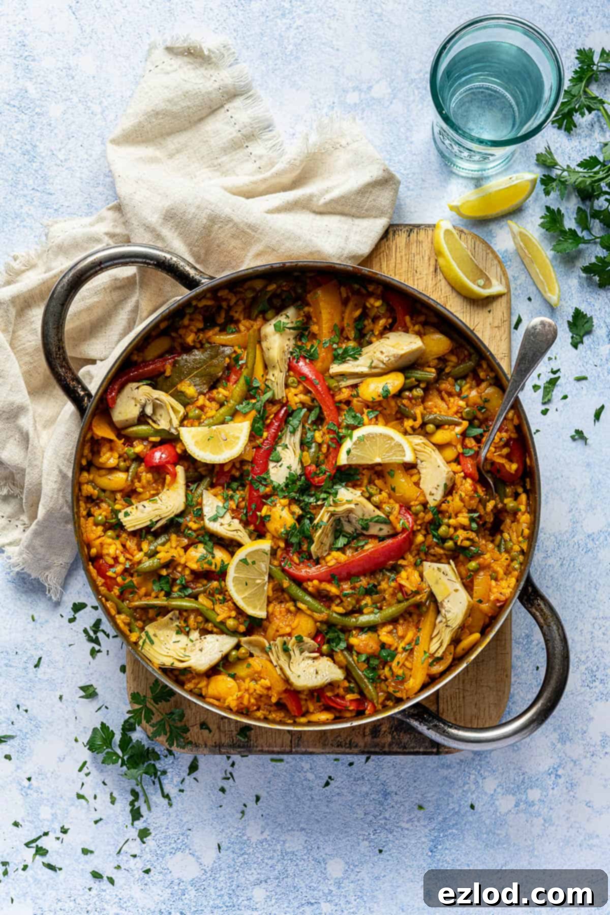 A pan of vegan vegetable paella on a wooden board on a blue background with a beige cloth, glass of water and fresh parsley.
