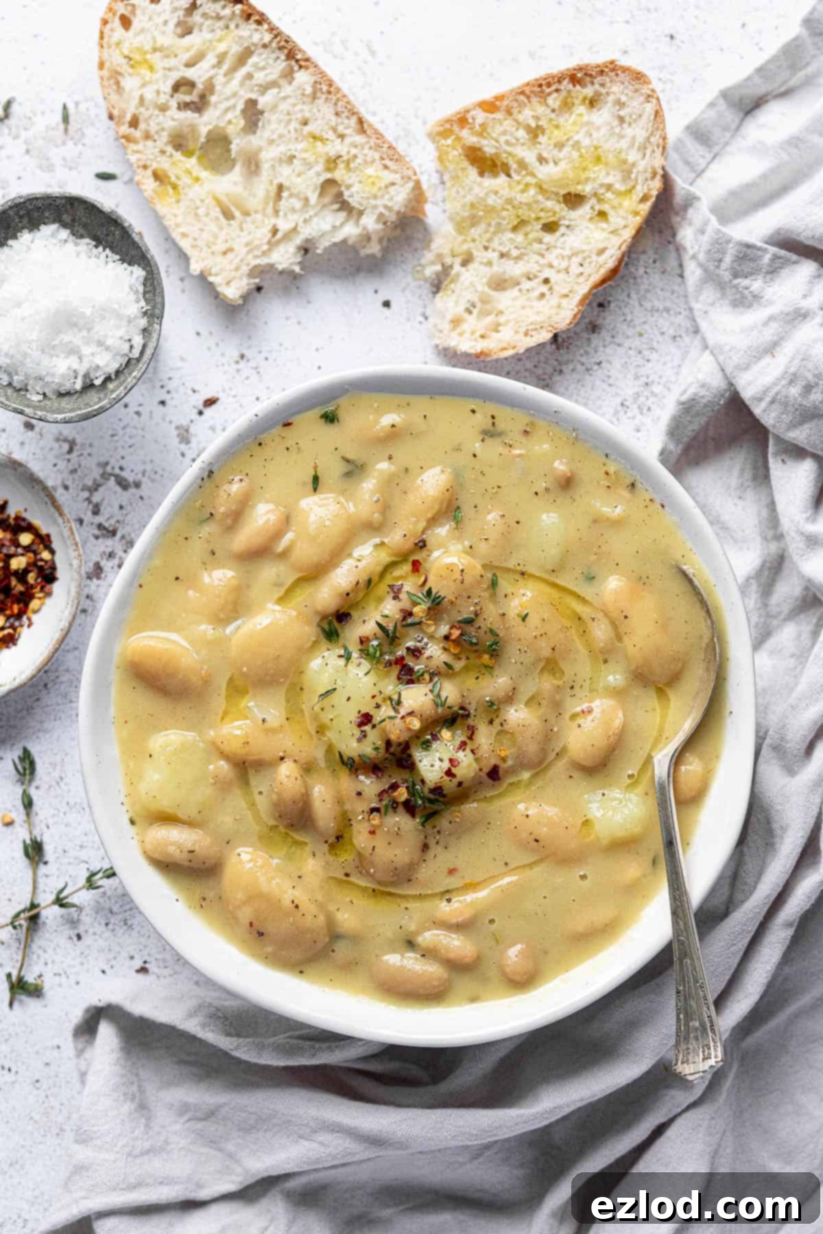 A bowl of rich, creamy white bean soup garnished with fresh thyme, served on a white background with a grey cloth, torn rustic bread, salt, and chilli flakes.
