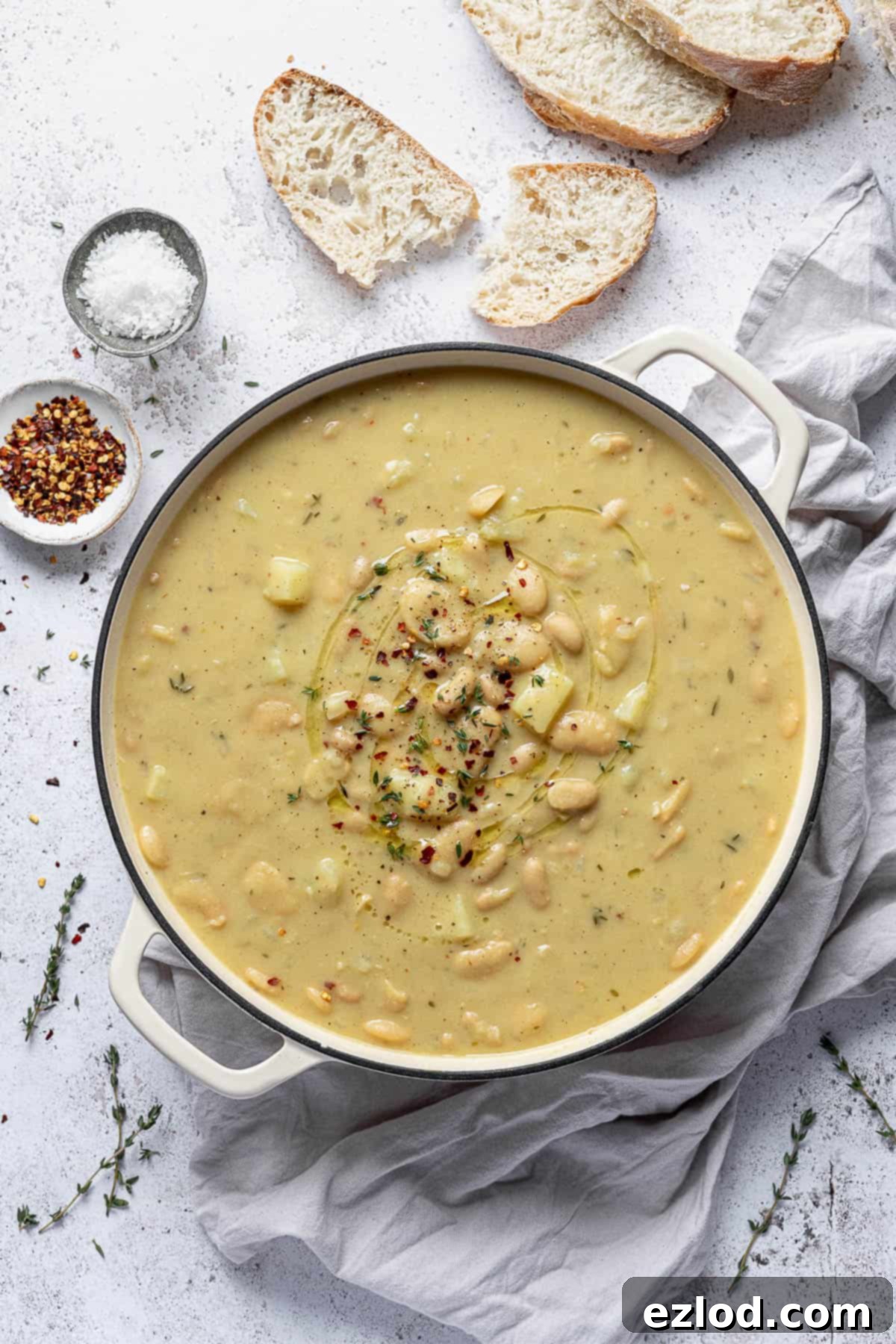 A large white pot filled to the brim with creamy white bean soup, artfully arranged on a white background with a contrasting grey cloth, accompanied by slices of rustic bread, small bowls of sea salt and red chilli flakes, and fresh thyme sprigs.