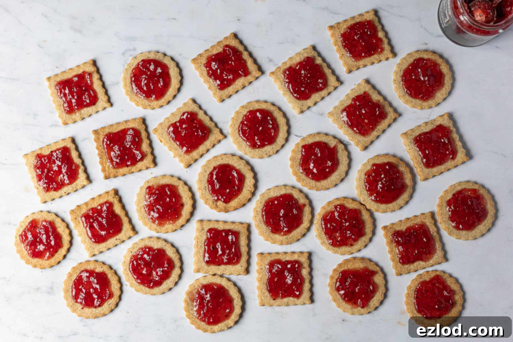 The bottom halves of vegan Linzer cookies, each generously spread with raspberry jam, waiting for their tops.