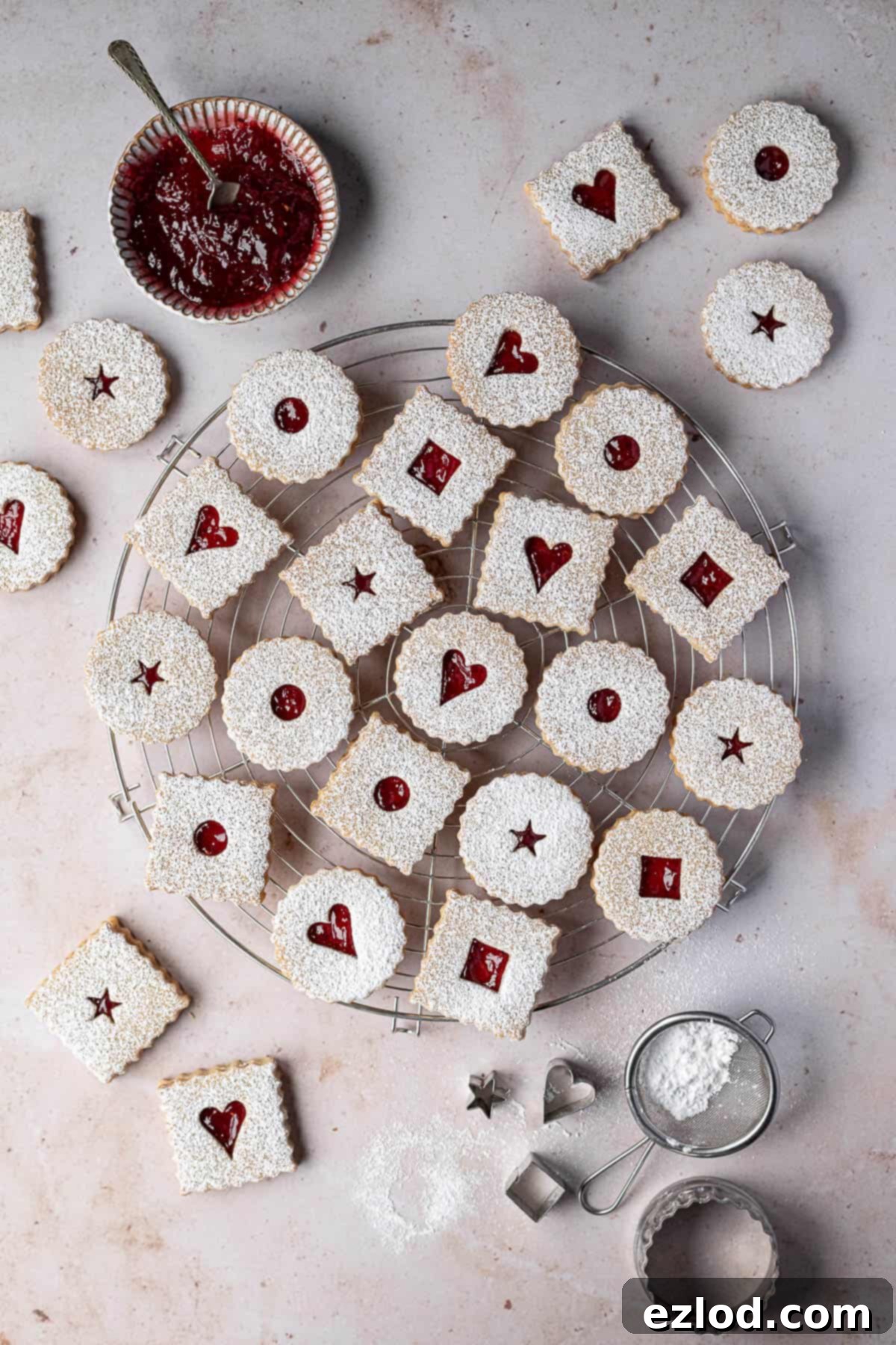 A beautiful arrangement of vegan Linzer cookies on a wire cooling rack against a marble background, with jam, icing sugar, and cookie cutters in the soft focus background.